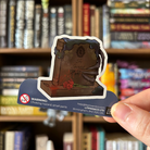 Book shaped sticker inspired by the fantasy genre, featuring a book designed to look like a tombstone, with creeping claws, blood, and carnations.