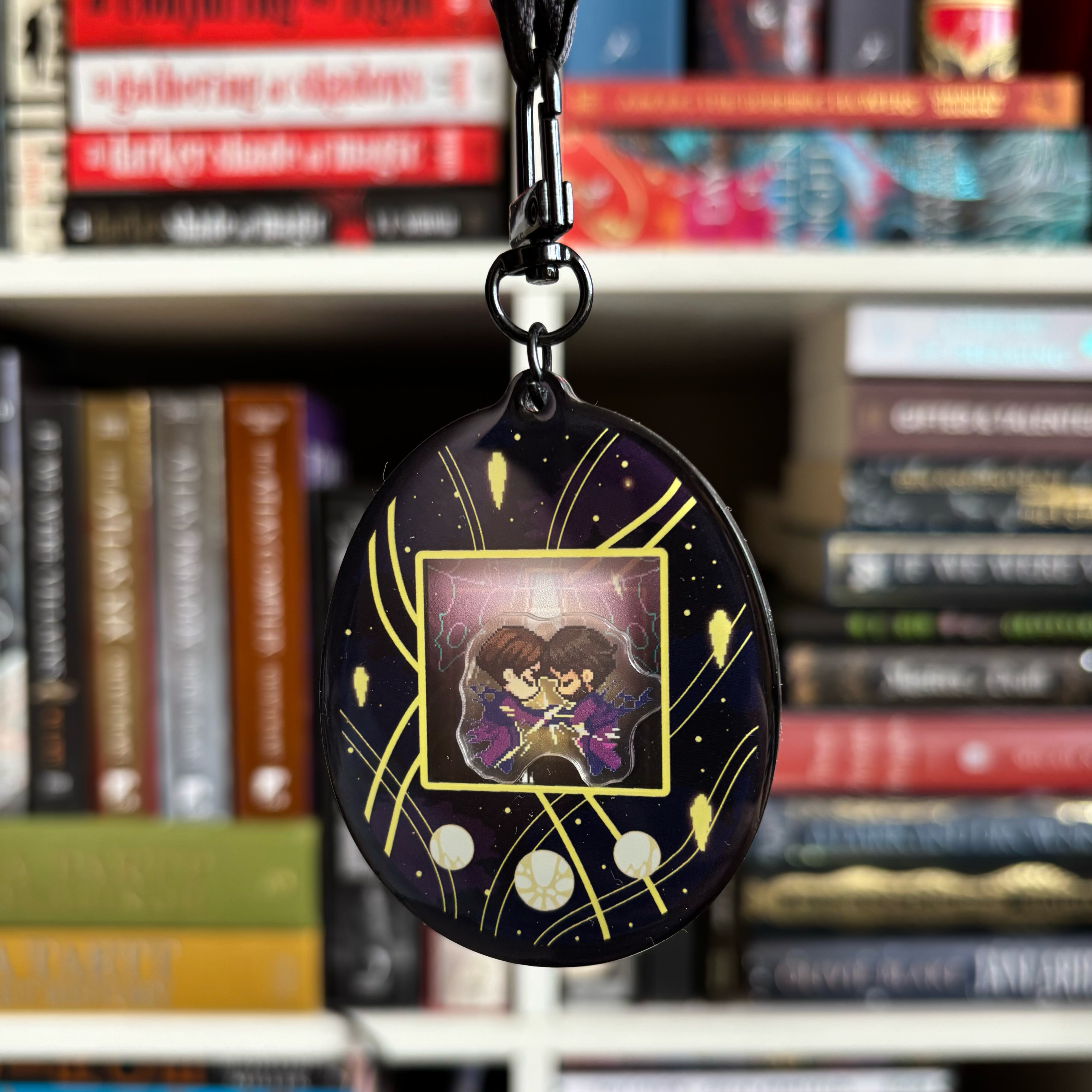 A black and gold Tamagotchi style shaker keychain inspired by Arcane, featuring Jayce and Viktor. Held in front of a book shelf.