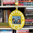 A yellow, white, and gold Tamagotchi style shaker keychain inspired by Arcane, featuring Jayce and Viktor. Held in front of a book shelf.