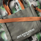 Grey canvas tote bag with white text reading "Ghost Hunting Supplies est. 1973" printed on the front. The bag has brown straps and is pictured against a background of page overlays.