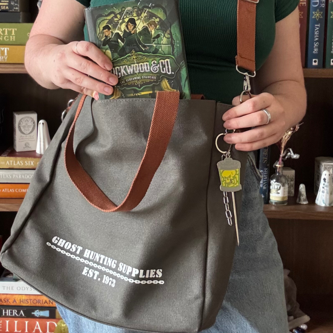 Grey canvas tote bag with white text reading "Ghost Hunting Supplies est. 1973" printed on the front. The bag has brown straps and is pictured being worn by a girl wearing a green t-shirt and blue jeans.