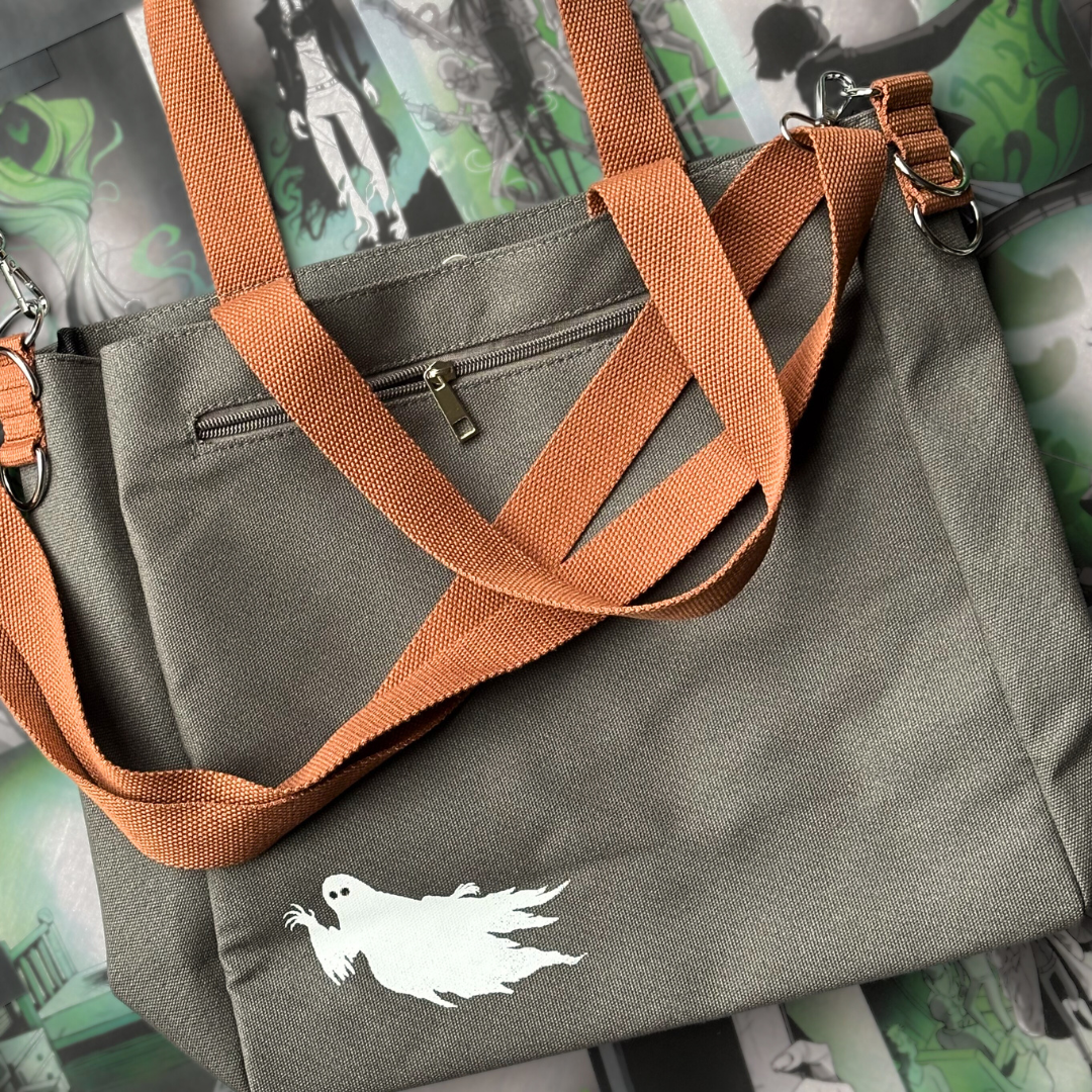 Grey canvas tote bag with a glow-in-the-dark white ghost printed in the bottom corner. The bag has brown straps and is pictured against a background of page overlays.