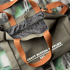 Grey canvas tote bag with white text reading "Ghost Hunting Supplies est. 1973" printed on the front. The bag has brown straps and a chain design inside, and is pictured against a background of page overlays.