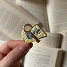 Officially licensed Murder Most Unladylike pin, featuring a book and a magnifying glass with the text "The Wells & Wong Detective Society," held against a background of blurred books.