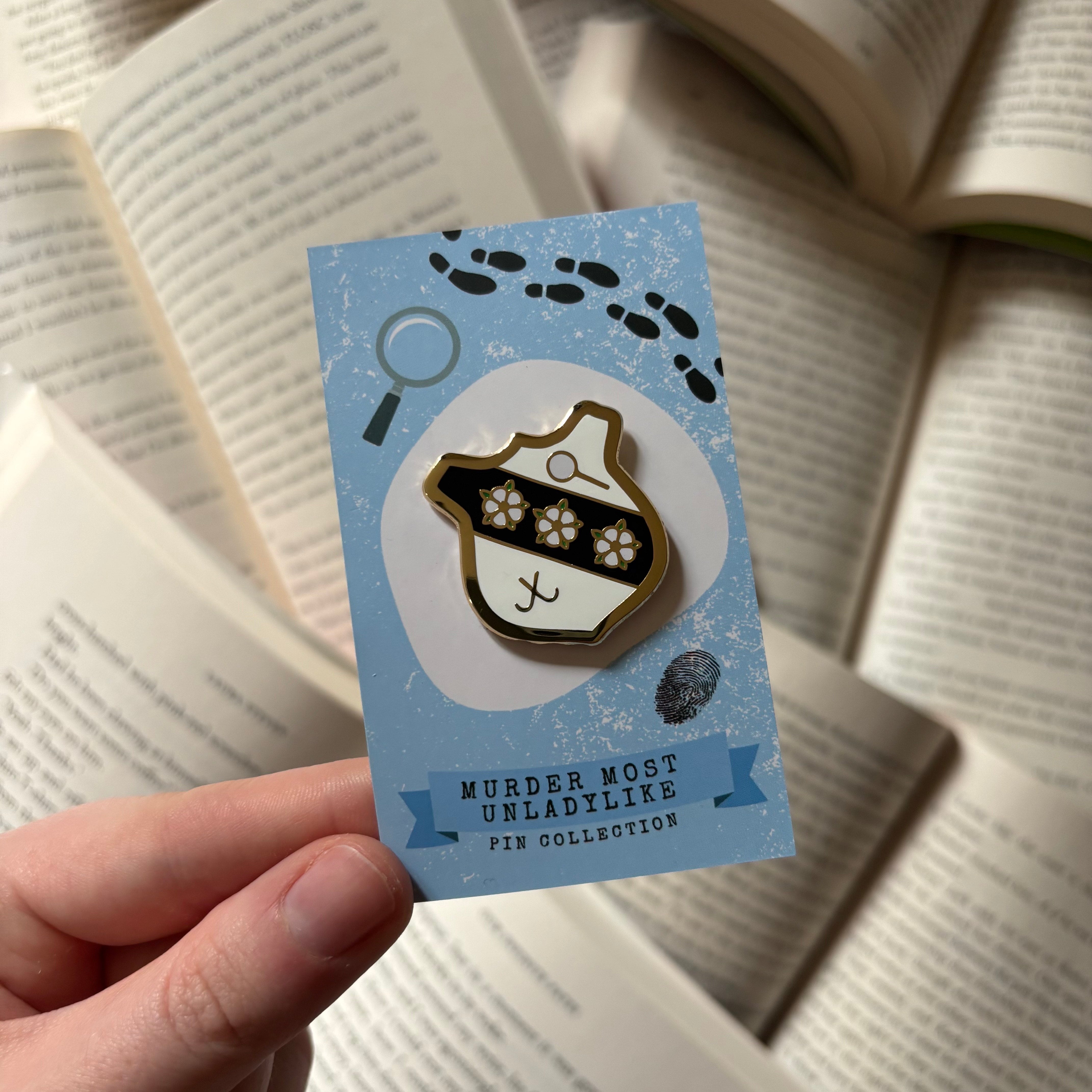 Official Murder Most Unladylike pin, featuring the crest of Deepdean school. Held above a background of blurred books