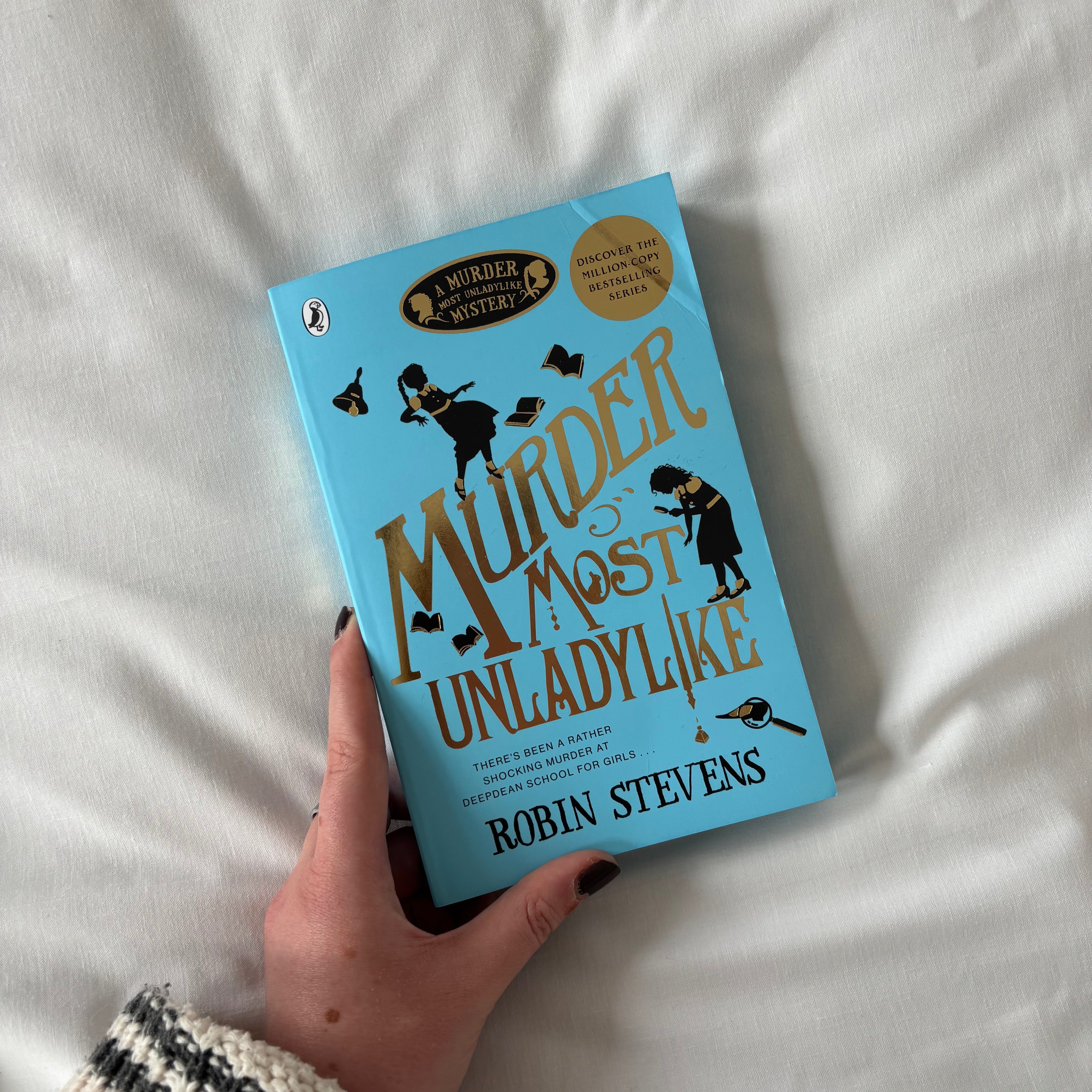 Book titled 'Murder Most Unladylike' by Robin Stevens held against a white fabric background