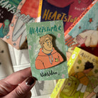 The Nick Nelson official enamel pin on a Heartstopper backing card, being held above a blurred background of Heartstopper books. 