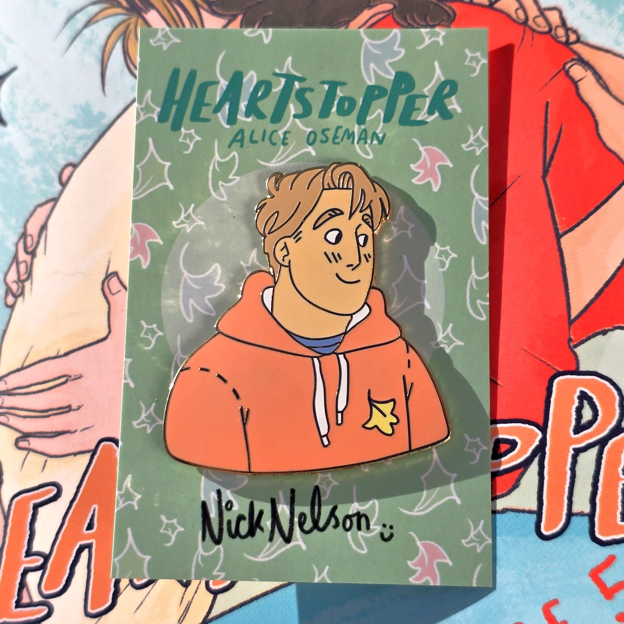 The Officially Licenced Heartstopper Nick Nelson Enamel Pin with backing card resting against a copy of Heartstopper. 