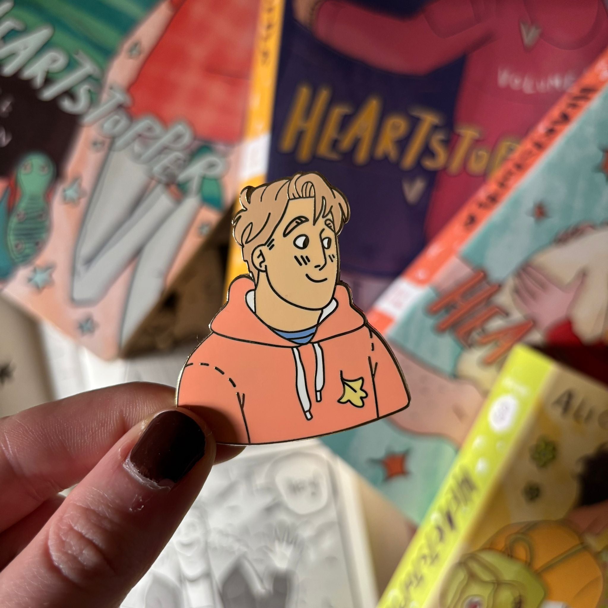 The Nick Nelson officiall licensed Heartstopper enamel pin being held above a blurred background of Heartstopper Graphic Novels.