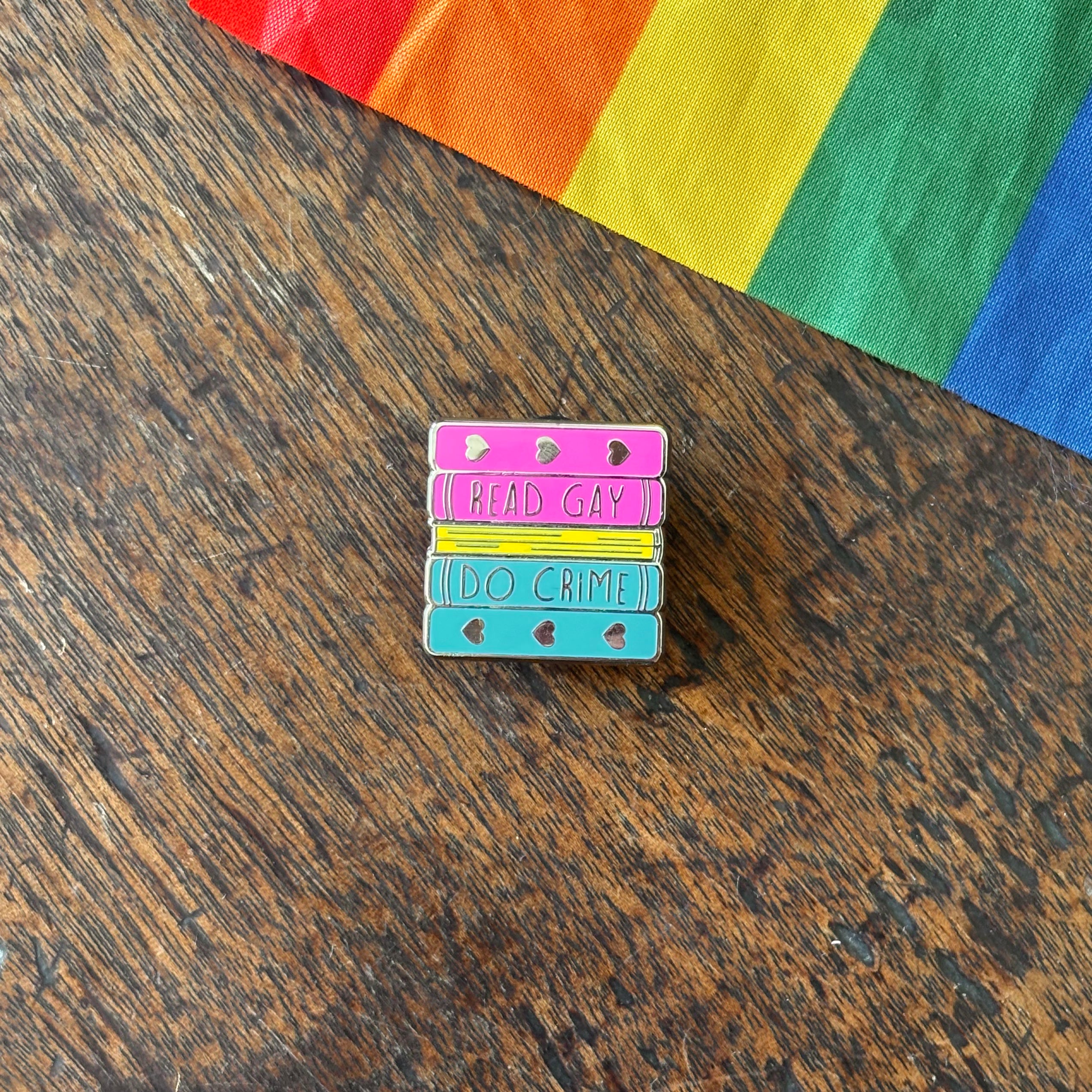 Pin with 'Read Gay, Do Crime' text on a wooden surface with a rainbow fabric background
