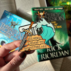 Percy Jackson inspired enamel pin, featuring a column, a sword, a wave, and the quote "The real world is where the monsters are," held above a background of books.