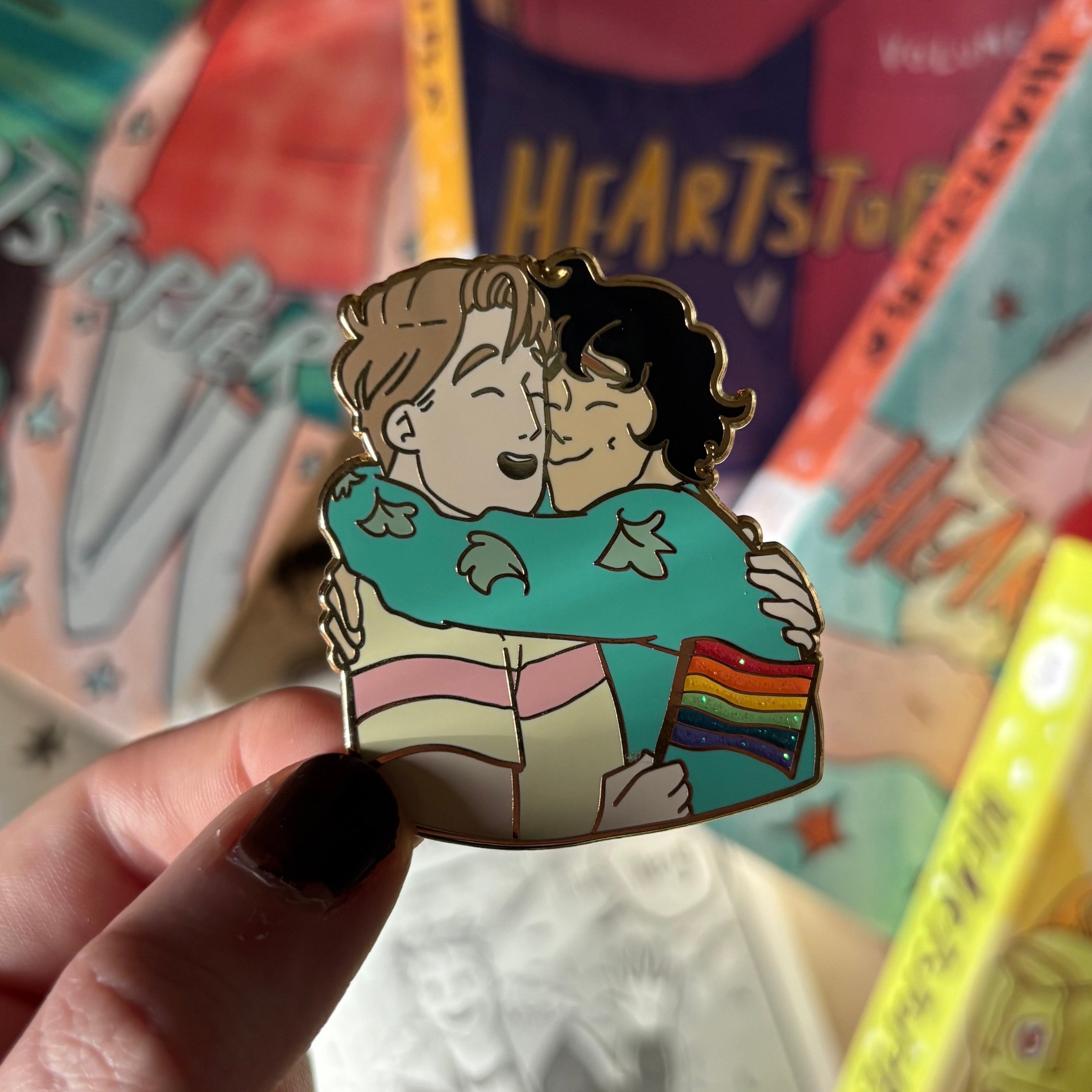 Officially licensed Heartstopper pride pin. Features Nick and Charlie in an embrace with Nick holding a Pride Flag., designed by Alice Oseman. Held against a background of Heartstopper graphic novels.