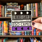 A large sticker designed to look like a stack of books in the colours of the asexual pride flag, with text reading "Read Gay Do Crime"