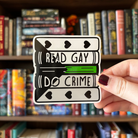 A large sticker designed to look like a stack of books in the colours of the demiromantic pride flag, with text reading "Read Gay Do Crime"