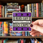 A large sticker designed to look like a stack of books in the colours of the greysexual pride flag, with text reading "Read Gay Do Crime"
