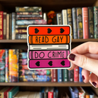 A large sticker designed to look like a stack of books in the colours of the lesbian pride flag, with text reading "Read Gay Do Crime"