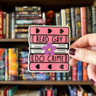 A large sticker designed to look like a stack of books in the colours of the sapphic pride flag, with text reading "Read Gay Do Crime"