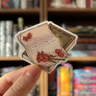 Book shaped sticker inspired by the romance genre, featuring a love letter, a rose, and a book of poems.