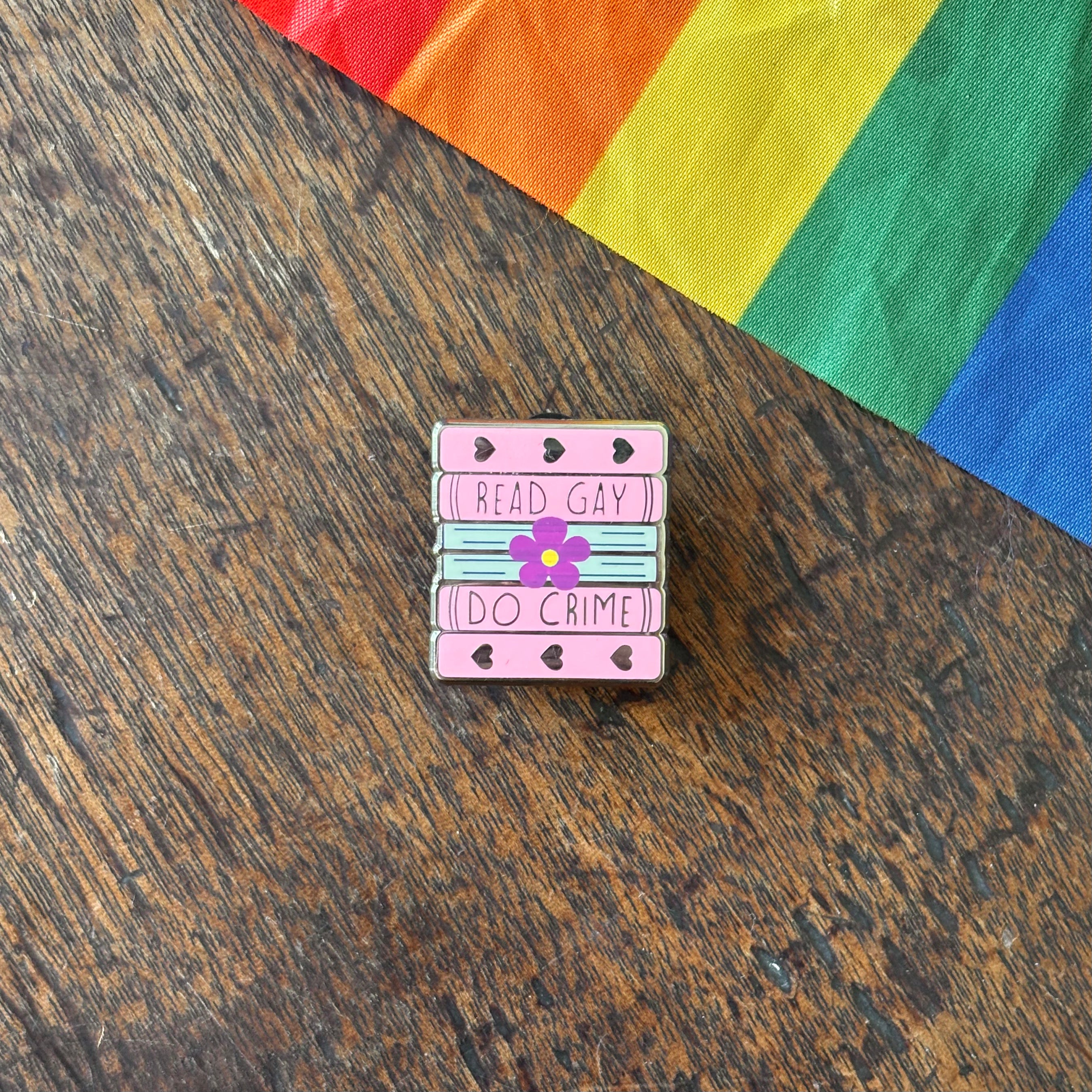 Pin with 'Read Gay, Do Crime' text on a wooden surface with a rainbow fabric background