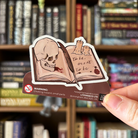 Book shaped sticker inspired byShakespeare plays, featuring a Shakespeare quote, a skull, some candles, and a quill on an open book.