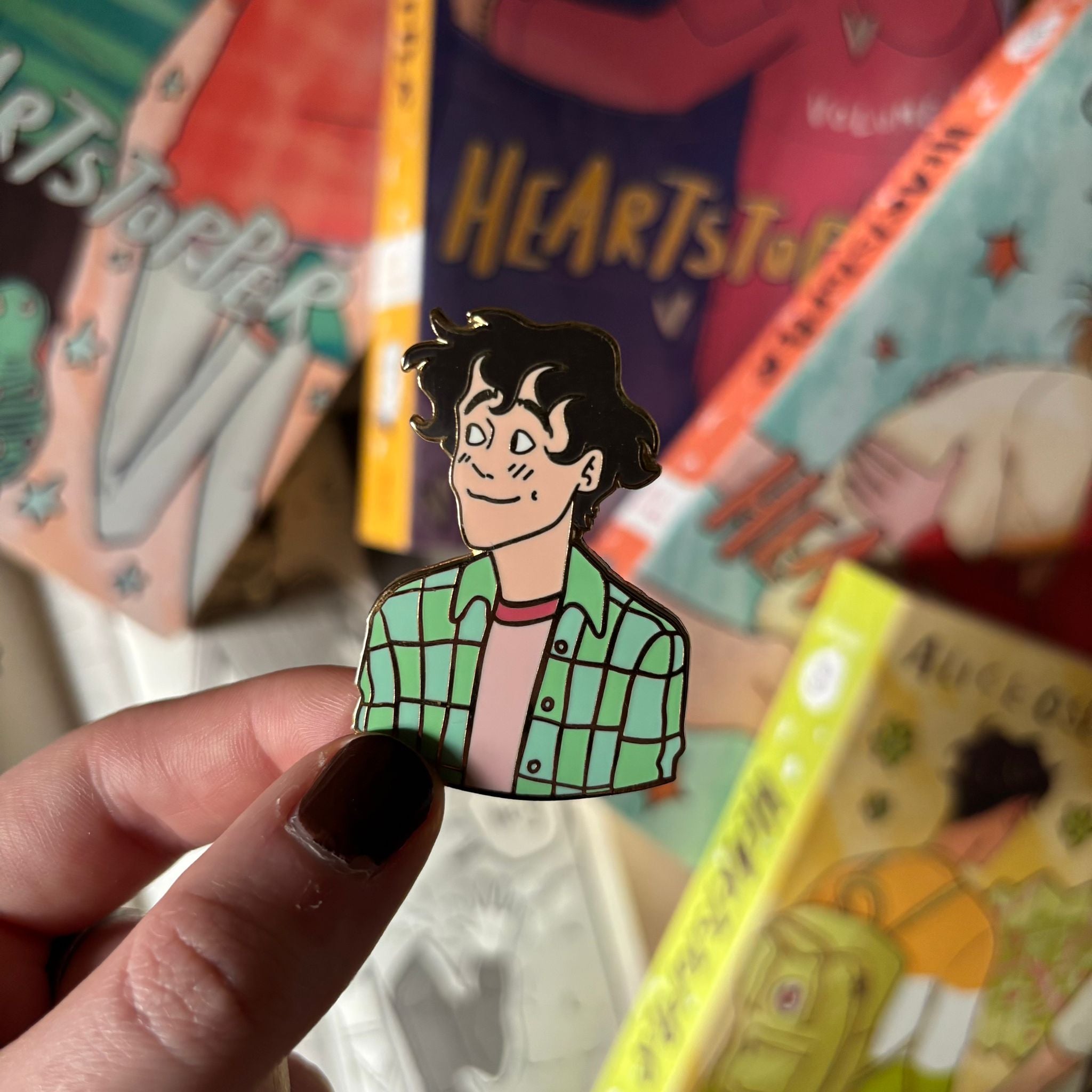 Heartstopper Charlie Spring enamel pin without backing card being held against a blurred background of Heartstopper Graphic Novels by Alice Oseman.