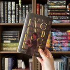 LitPins&Co special edition of The Incandescent, held in front of a bookshelf.