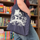 A woman stood in front of a bookshelf, wearing a burgundy top, blue jeans, and a navy tote bag. The tote bag features a school crest of the fictional school "Wickwood Academy" from CG Drews' Don't Let The Forest In. The crest features a book, a dove, a tree, and a graduation hat, with the school name along the bottom surrounded by leaves.