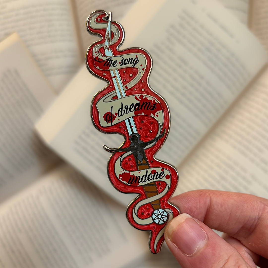 Enamel pin inspired by Ashdrinker from Empire of the Vampire by Jay Kristoff, held against a background of open books.