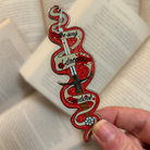 Enamel pin inspired by Ashdrinker from Empire of the Vampire by Jay Kristoff, held against a background of open books.