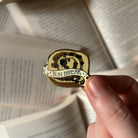 An enamel pin shaped like a bun, with the text "bun break," held above a background of open books