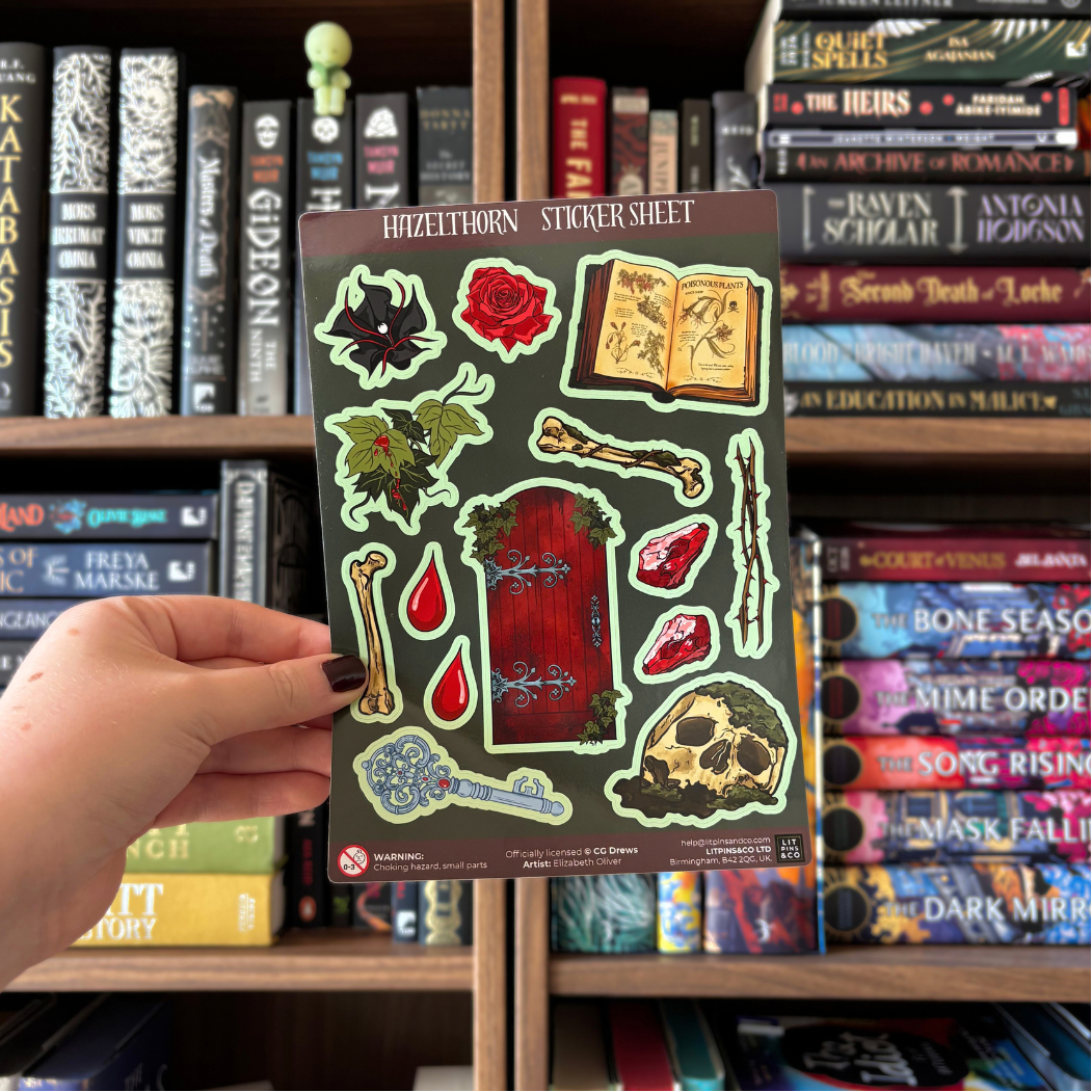 A gothic-themed sticker sheet titled 'HAZELTHORN STICKER SHEET' featuring illustrations of a red door, skull, bones, rose, key, and other mystical motifs on a dark green background. Sticker sheet is held against a blurred bookshelf