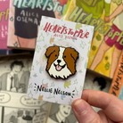Nellie Nelson enamel pin inspired by Alice Oseman's Heartstopper series. The pin is being held in front of the Hearstopper graphic novels.
