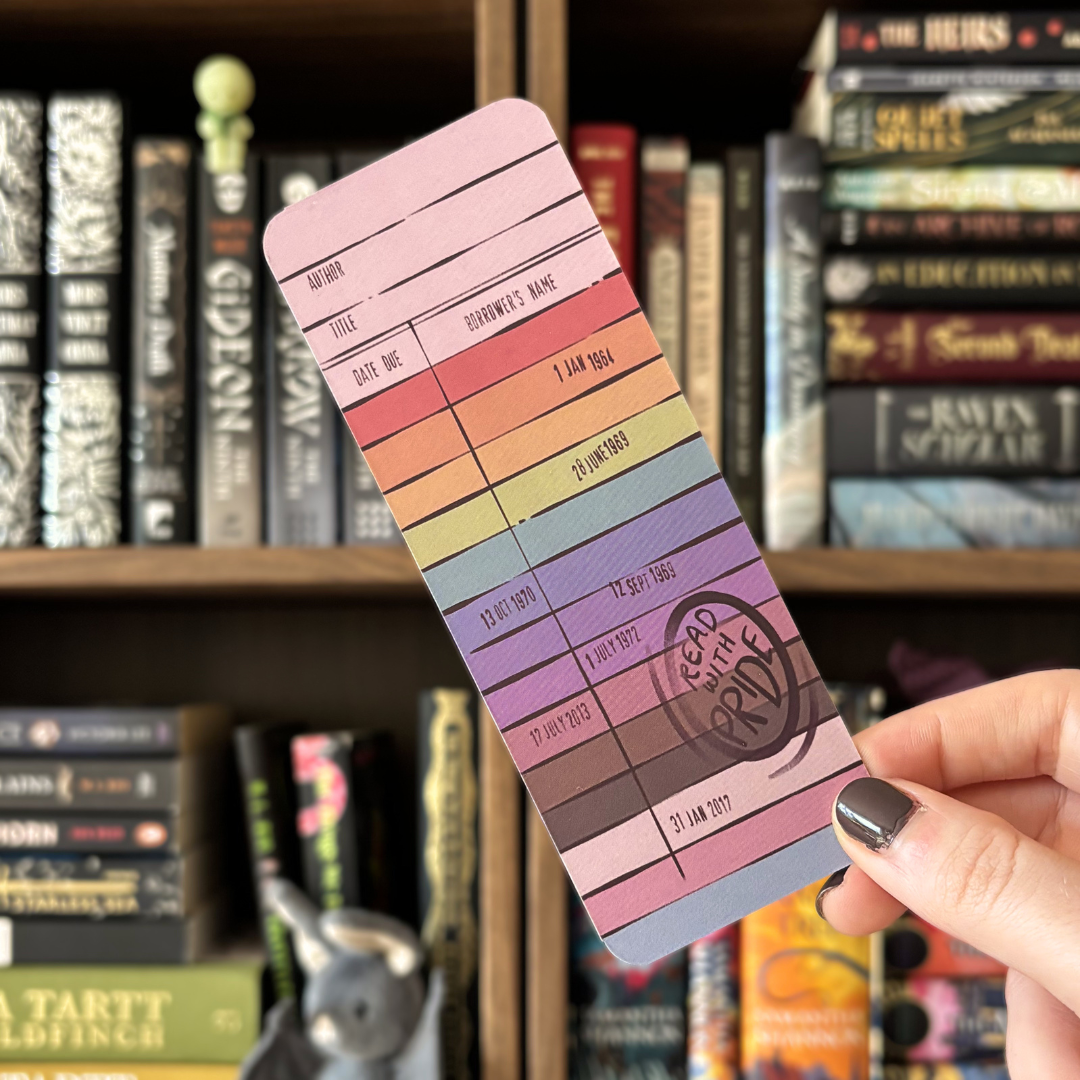 Read with pride bookmark, designed to look like a library borrowing slip and coloured in the colours of the progress pride flag. Bookmark is held in front of a blurred bookshelf.