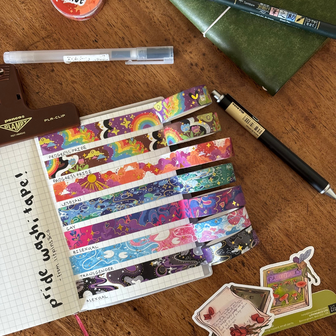 A set of colourful washi tapes inspired by various pride flags, displayed on a grid notepad and surrounded by various pieces of stationary