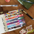 A set of colourful washi tapes inspired by various pride flags, displayed on a grid notepad and surrounded by various pieces of stationary
