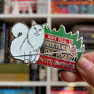 Enamel pin inspired by Margaret Rogerson's Sorcery of Thorns, featuring a cat and sword design and text reading "Not all battles are fought with swords" held in front of a bookshelf.