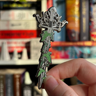 Sword pin inspired by the fantasy world of Middle Earth, held against a blurred background of bookshelves.
