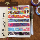 A set of colourful washi tapes inspired by various pride flags, displayed on a grid notepad and surrounded by various pieces of stationary