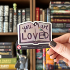 Book-shaped sticker with the text "you are LOVED" decorated in colours of various pride flags