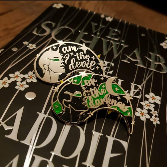 Am I the Devil or The Darkness? Dual enamel pins resting on top of The Invisible Life of Addie LaRue by V.E Schwab