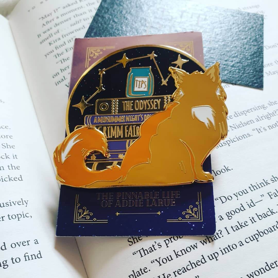 Books Feed The Mind, Tips Feed The Cat enamel pin laying against an open copy of The Invisible Life of Addie LaRue by V.E Schwab